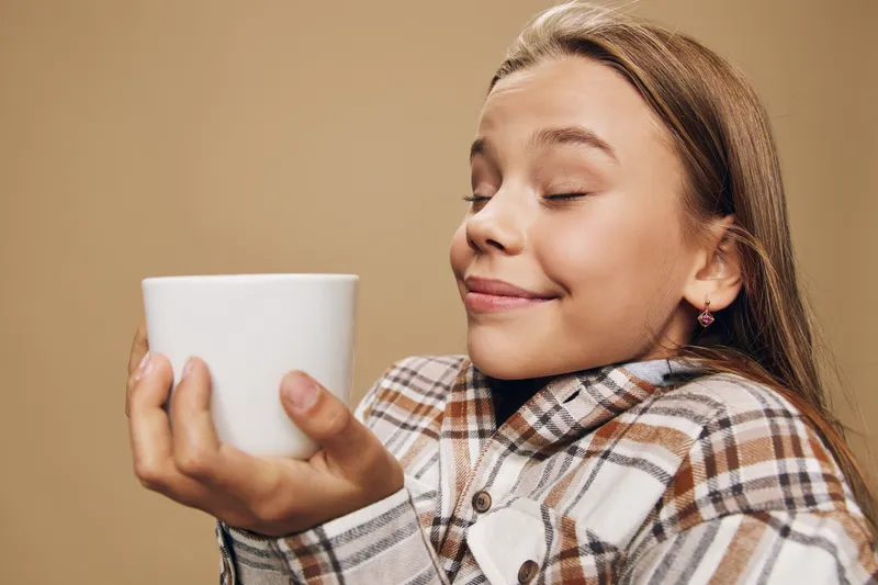 Young girl enjoying hot beverage with closed eyes and smile holding white cup wearing cozy plaid shirt against beige background indoors. Relaxation, comfort, warmth and happiness.