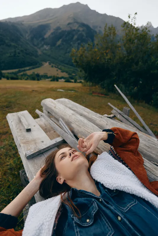 Woman portrait outdoors lying on wooden bench in meadow near mountains, relaxing with eyes closed and hand on forehead, wearing denim shirt and shearling jacket in natural landscape.