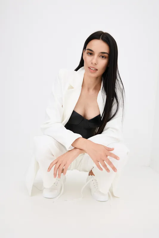 Young woman crouching in stylish white suit and black top, with long dark hair and minimal makeup, posing against plain white background in bright light.