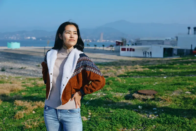 Woman in denim jeans and a warm jacket stands in a green field under a clear sky, showcasing casual outdoor fashion and a confident pose in natural light.