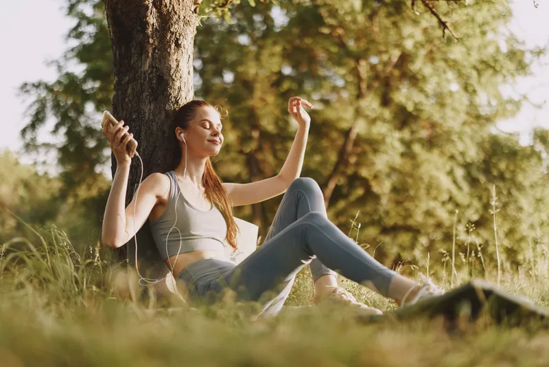 Young woman enjoying music outdoors, sitting under a tree, dressed in casual sportswear, exuding joy and relaxation in a sunny, green environment