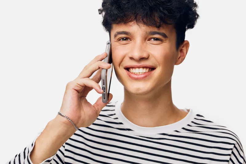 Young man smiling while talking on smartphone, wearing striped shirt with curly hair against plain white background, casual and cheerful atmosphere.