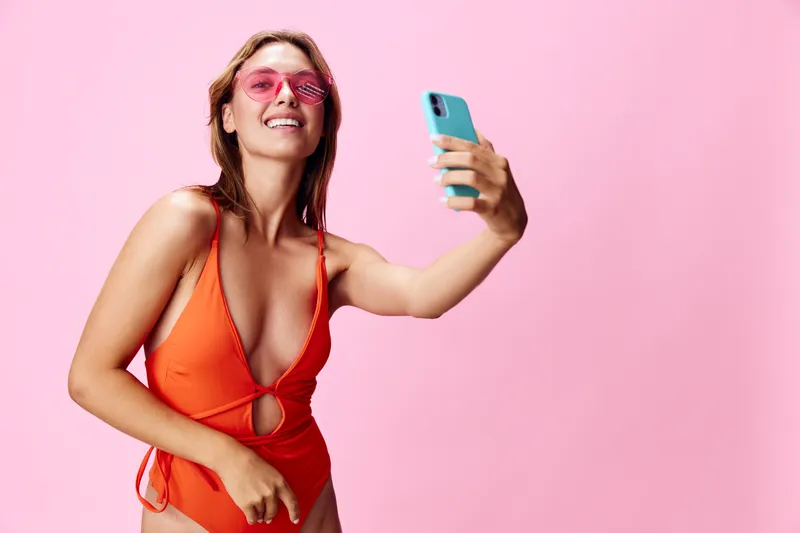 Яркая женщина в оранжевом купальнике делает селфи на телефон на красивом розовом фоне