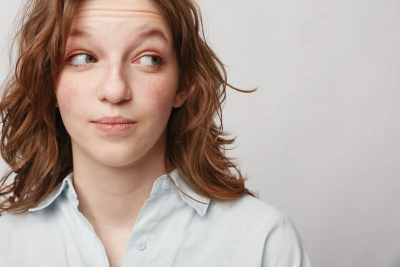 Young woman with wavy brown hair looks sideways with a thoughtful expression wearing a light blue shirt against a plain background. Concept of curiosity and contemplation.