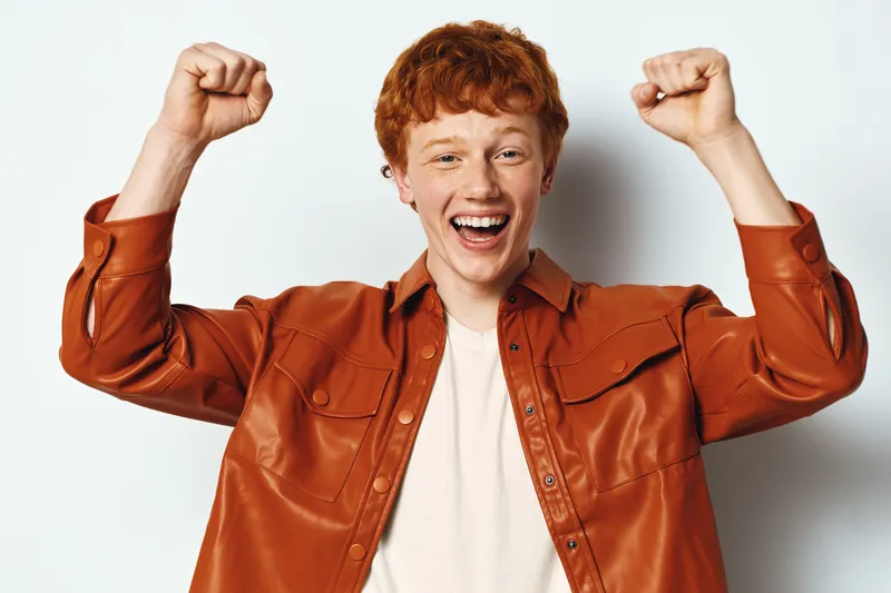 Young man with red hair wearing brown leather jacket smiling and raising fists in celebration or excitement on white background. Happy male portrait with positive emotions.