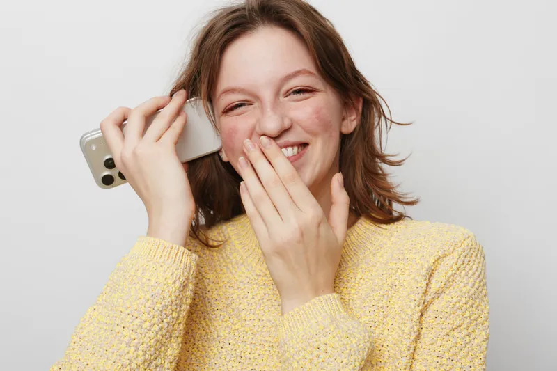 Smiling young woman in yellow sweater holds smartphone near ear and covers mouth with hand. Casual lifestyle, communication, happiness, and joyful expression on light background.