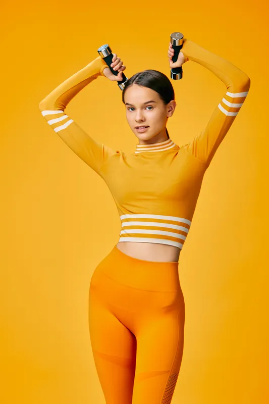 Fitness woman in bright orange athletic wear raises dumbbells overhead during a high-energy workout, showcasing strength and balance against a vivid yellow backdrop in a bold fitness scene.