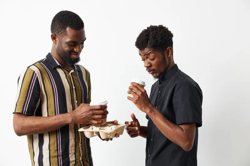 Two smiling African American men sharing a moment with coffee cups, enjoying each other’s company in a bright, neutral-toned atmosphere, creating a warm vibe.