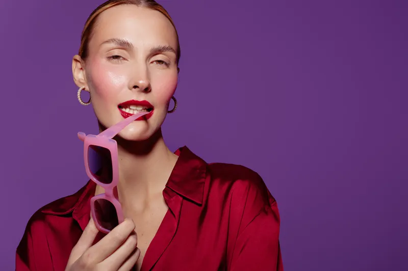 Young woman in red blouse holds pink sunglasses near lips on purple background. Fashion portrait with bright makeup and confident expression. Concept of style and beauty.