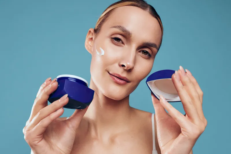 Woman skincare beauty cosmetic cream moisturizer face holding jar container, young smiling model in studio, holding blue jar and white container, concept skin care daily routine.
