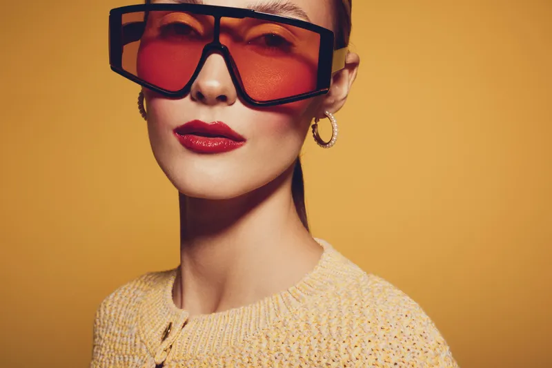 Stylish woman with red lipstick and large orange sunglasses poses against yellow background. Fashion portrait with trendy accessories and confident expression in warm studio lighting.
