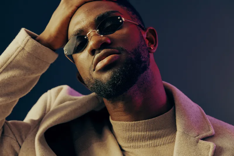 Stylish young man in sunglasses and a beige coat posing confidently against a dark blue and purple background, exuding a modern and trendy vibe