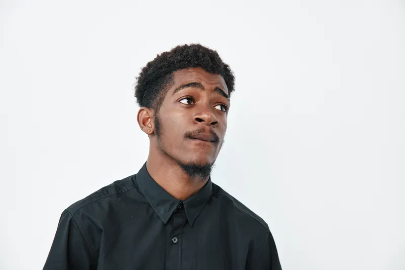 Young black man with dark skin wearing black shirt looking sideways on white background. Portrait of African ethnicity male with short curly hair and thoughtful expression in studio.