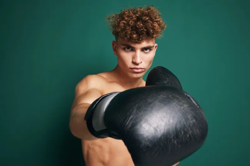 Young male boxer with curly hair wearing black boxing gloves in fighting stance against green background. Athletic shirtless man training for boxing or martial arts workout.
