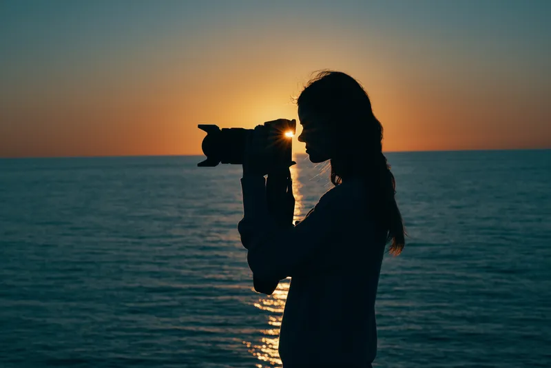 Силуэт женщины-фотографа на закате у моря, вид сбоку