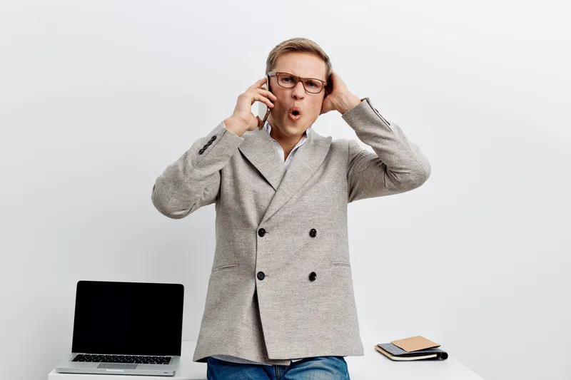 Confused man wearing glasses and a gray coat expressing surprise, standing against a plain light background with a laptop and notepad on the table, studio portrait. People lifestyle concept
