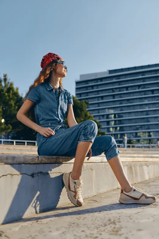 Lifestyle woman in modern boho-western denim street style wearing a red bandana and sneakers, sitting outdoors under bright natural sunlight in film color tones.