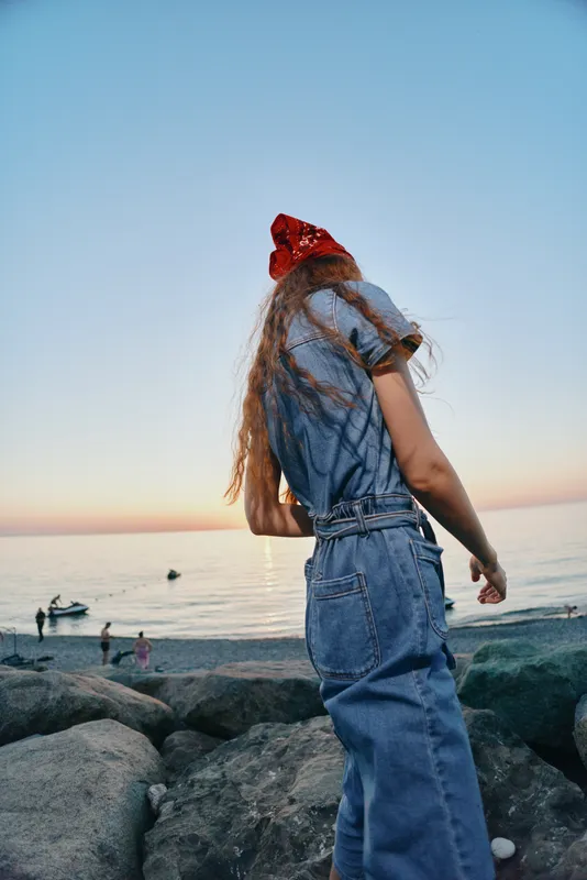young woman wearing denim outfit and red bandana standing on rocky shore near calm sea at sunset with clear sky and peaceful mood