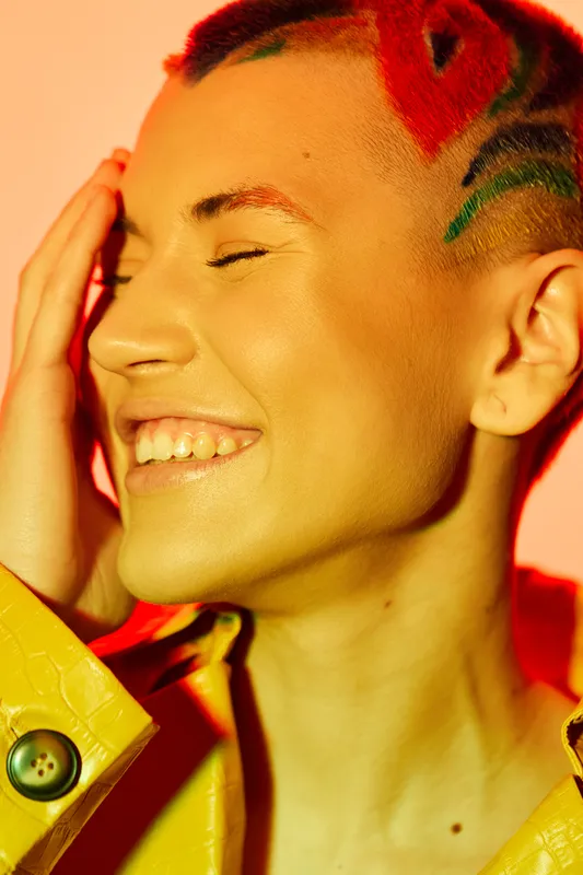 Smiling woman with colorful hair and bright makeup wears yellow jacket, expressing joy and vibrant energy in warm lighting, close-up portrait.
