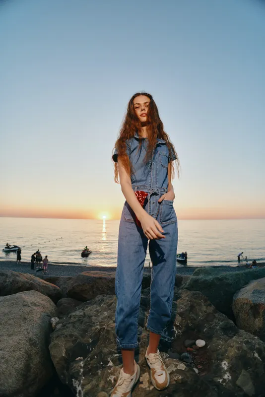 lifestyle woman in modern boho-western denim street style standing on rocky shore at sunset, film color with casual trendy outfit and relaxed pose