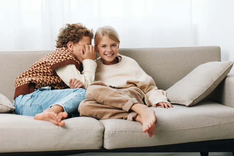 Two children sit close on a cozy sofa, one whispering softly to the other, both smiling and relaxed, capturing a candid moment of friendship and playful secrecy.