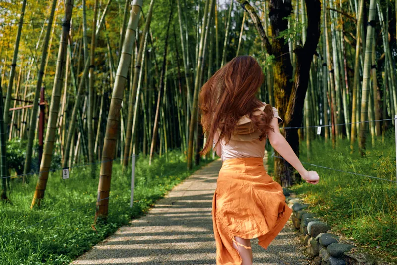 woman running on a bamboo path in nature, back view of a girl in a flowing orange dress on a sunlit trail, motion and carefree summer outdoor scene with green forest surroundings