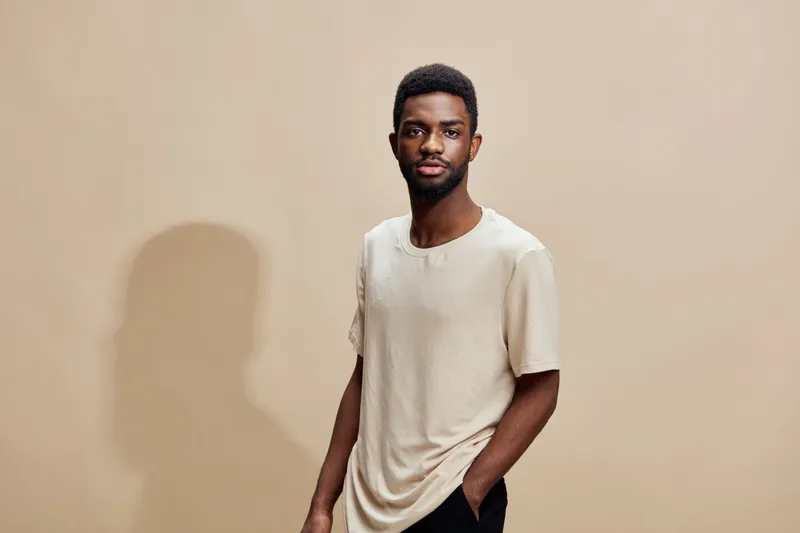 Young African man in a casual beige shirt with a confident expression, standing against a neutral background, embodying modern style and self assurance