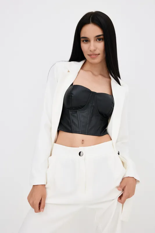 Young woman in white suit and black corset posing confidently with hands in pockets, displaying stylish fashion look and modern elegance on plain background.