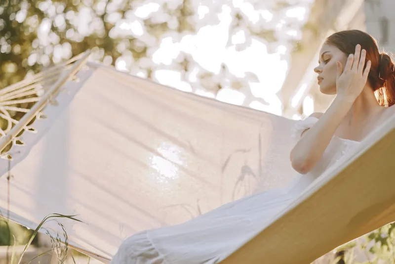 Serene young woman lounging in a hammock during a sunny day, wearing a light white dress, enjoying a peaceful moment in nature