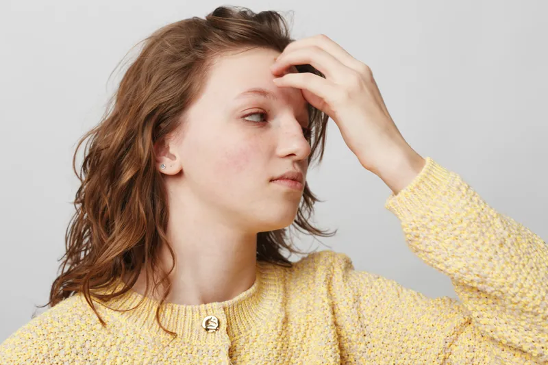 Young woman with curly hair in yellow sweater touches forehead, looking away with thoughtful expression. Studio portrait on light background, concept of contemplation and casual fashion.