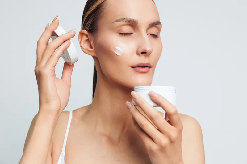 Young woman with blonde hair applies white face cream on her cheek while holding an open jar of cream against a light gray background in a skincare routine.