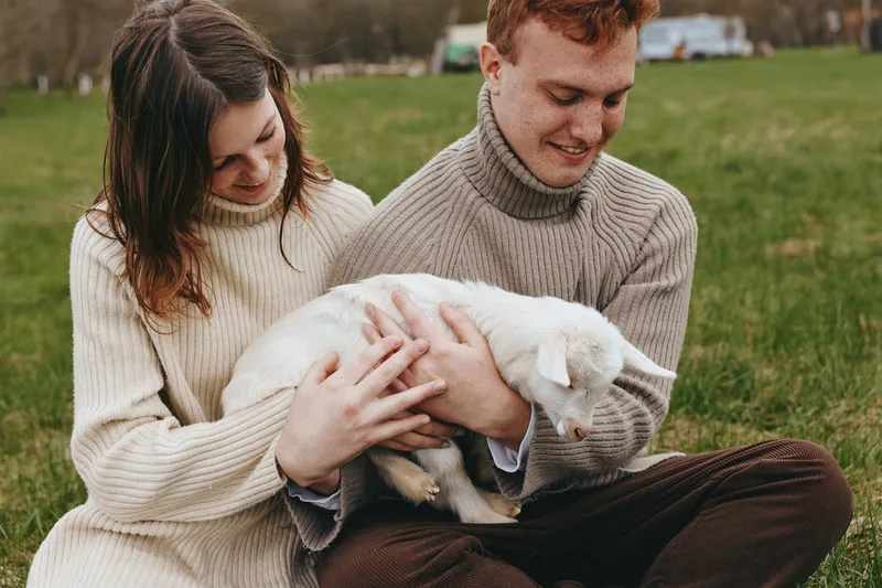Young couple in cozy sweaters sits on grass holding a white baby goat enjoying a calm moment in a natural outdoor setting during autumn.