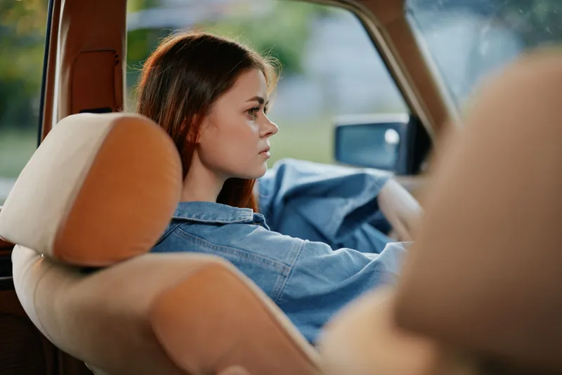 Young woman in denim jacket sitting in a car, reflecting on life with a serene expression, warm daylight highlighting her features, summer vibe.
