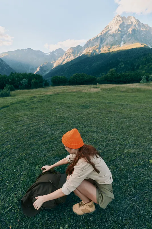 A young caucasian woman wearing an orange beanie crouches on a grassy field, arranging a backpack while rugged mountains loom in the distance, as the scene conveys preparation and calm.