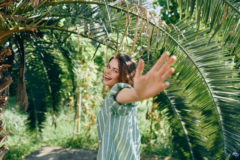 Joyful woman reaches toward the camera in a sunlit tropical garden, surrounded by lush palm fronds and vibrant greenery.