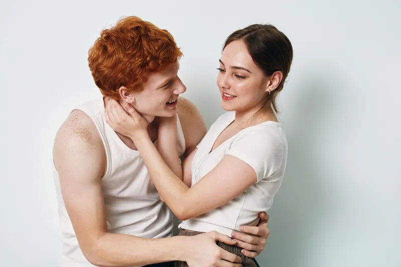 Young couple smiling and embracing while looking at each other in casual white clothes isolated on white background, romance, love and happy relationship concept