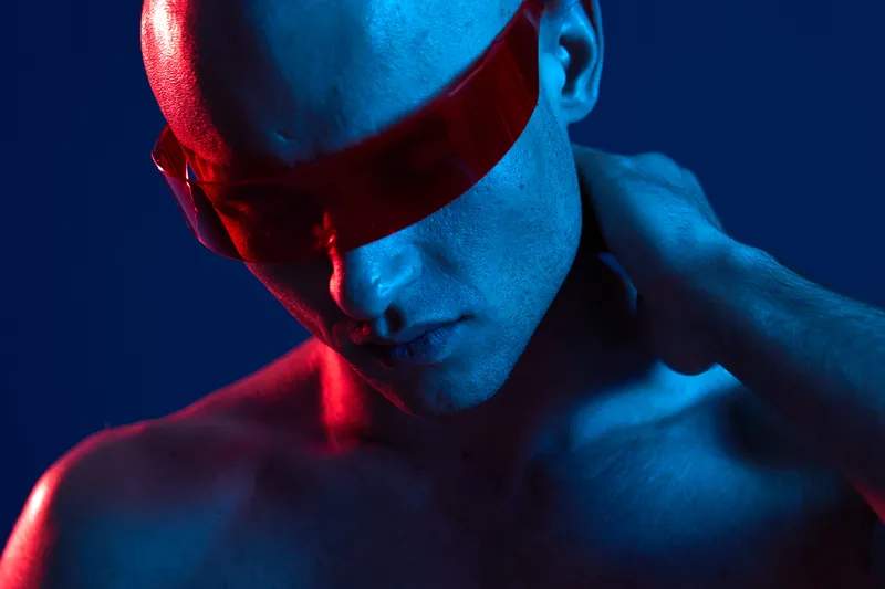 Stylish man with futuristic glasses, highlighted in vibrant blue and red lighting, showcasing a contemplative mood in an artistic black and white portrait.