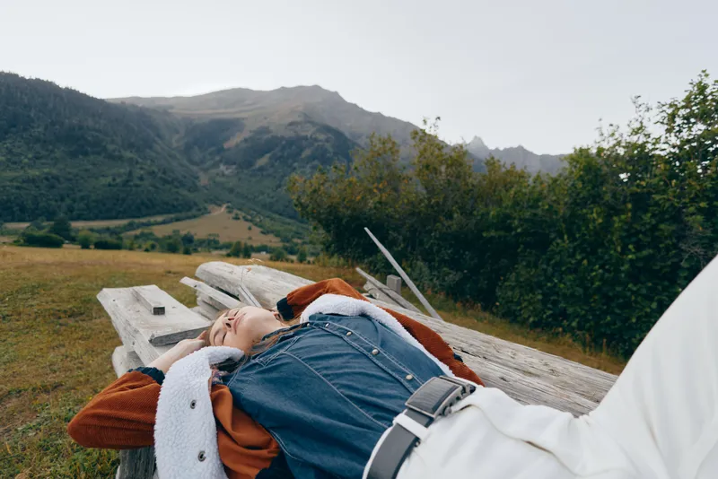 Woman lying on wooden bench in meadow near mountains, relaxing in nature during travel. Casual denim jacket and white pants, peaceful countryside rest and scenic landscape escape.