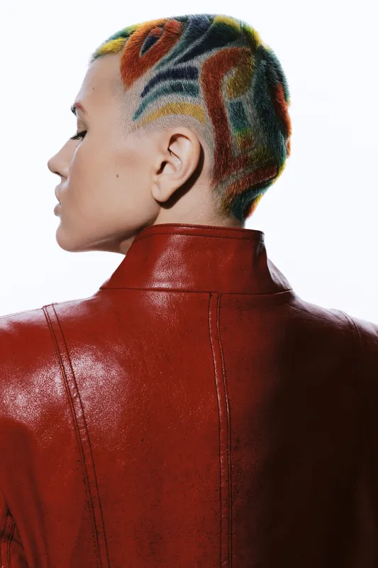 Woman with colorful shaved hair wearing a red leather jacket, showcasing bold hairstyle and edgy fashion against a clean white background.