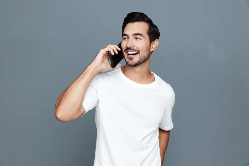 Smiling young man wearing white t-shirt talks on mobile phone with cheerful expression, standing against a solid gray background. Lifestyle and communication concept.