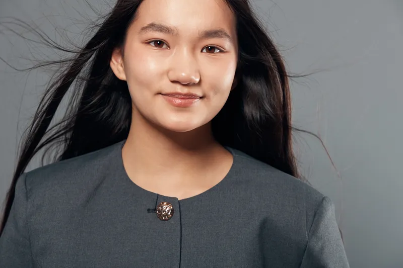 Young woman with long black hair and natural makeup wearing gray formal jacket looking confidently at camera with subtle smile on neutral gray background with studio lighting.