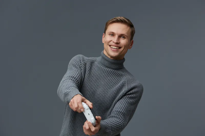 Smiling man in gray turtleneck sweater holding gaming controller, looking joyful, casual indoor setting, positive mood, lifestyle portrait, happiness concept.