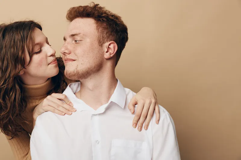Young couple in casual clothes embraces tenderly, looking at each other with affection. Warm beige background creates intimate and loving atmosphere. Concept of romance and connection.