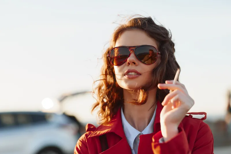 Confident woman wearing sunglasses and red jacket outdoors holding cigarette with blurred car background during golden hour with warm sunlight highlighting her face and hair