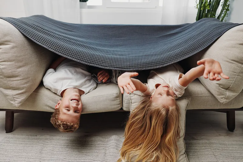 Two happy children lying upside down on a couch covered with a blanket, smiling and playing in a cozy living room with natural light and plants visible.
