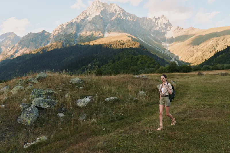 Young woman with backpack hikes through a mountain meadow surrounded by rocky peaks under a partly cloudy sky, enjoying an active outdoor lifestyle.