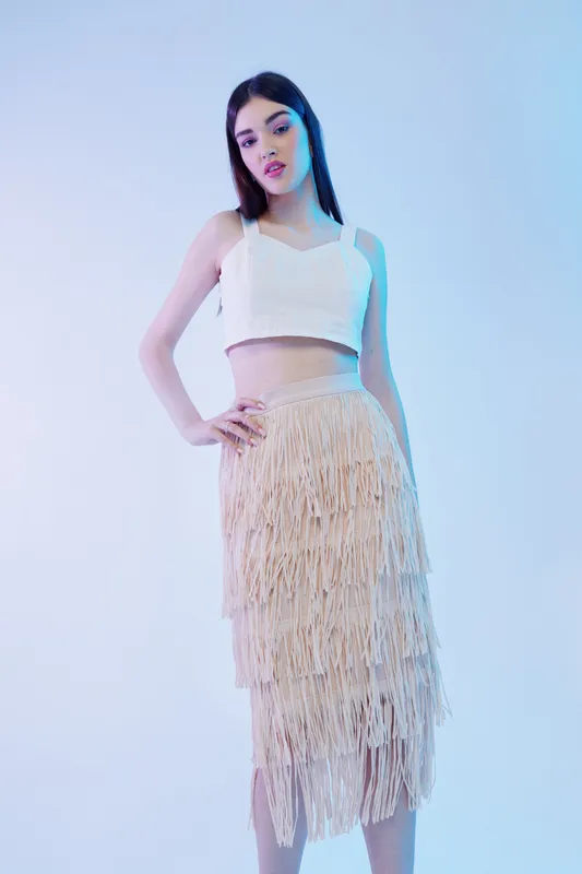 Young woman in a white crop top and fringed skirt poses confidently against a light background. Concept of woman beauty fashion, elegance, and modern style.