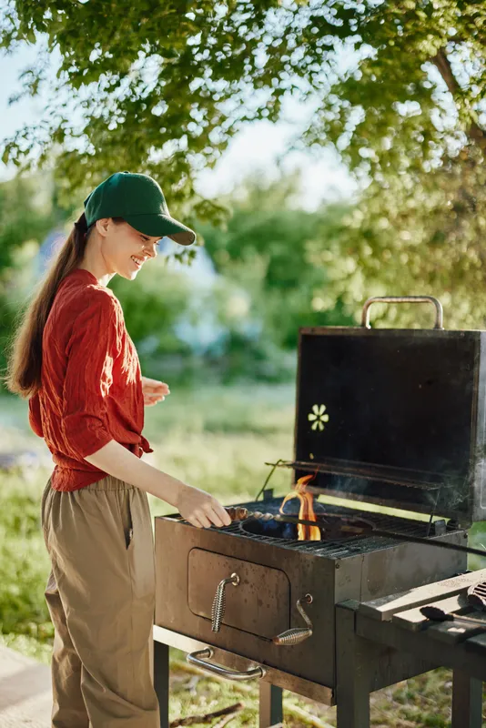 Young woman grilling outdoors in a casual outfit, smiling while enjoying a sunny day, surrounded by greenery and cooking delicious food on a barbecue