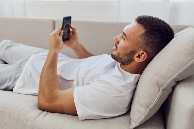 Relaxed man using smartphone while lounging on a cozy sofa, enjoying leisure time at home. Comfortable setting for a modern lifestyle in 2025.