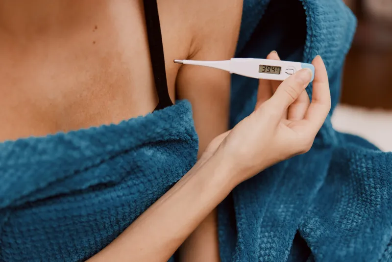 Женщина, укутанная в синее одеяло, в бюстгальтере и топе-майке, использует электронный термометр